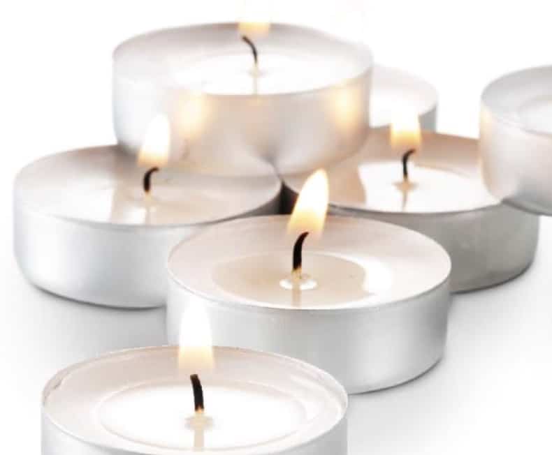 Several lit tealight candles with white wax in small, round, silver metal holders are clustered together on a white surface near a delicate air dry clay house. The flames flicker gently, casting a warm, soft glow around the charming miniature structure.